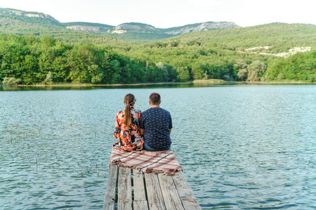 The couple sits on a wooden pier on the lake. There is a green forest and mountains around the lake....