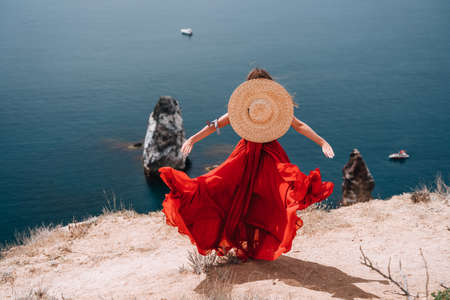 A woman is seen from behind in a Red flying dress fluttering in the wind. In a straw hat, arms outstretched against the sea with rocks and boats. The concept of romance, travel and tourism.の写真素材