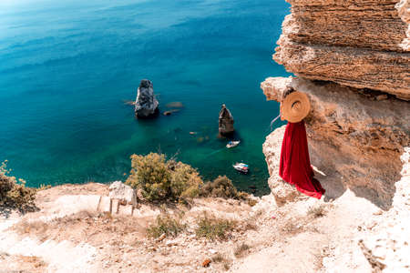 From behind, a woman is seen in a red flying dress fluttering in the wind. In a straw hat, walking down the stairs against the background of the sea with rocks and boats. The concept of travelの写真素材