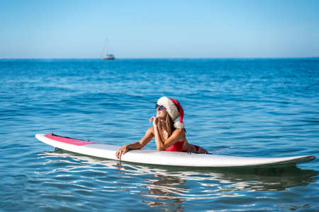 A woman in a red bikini and a Santa hat lies on a SUP board with the sea and a yacht floating on the horizon. He looks away and smiles.の写真素材