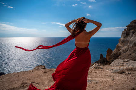 From behind, a woman is seen in a red flying dress fluttering in the wind. In a straw hat, walking down the stairs against the background of the sea with rocks. The concept of travelの写真素材