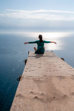 A girl sits on a wooden springboard for jumping with a rope. In a dark green dress with her hands in the air.の写真素材