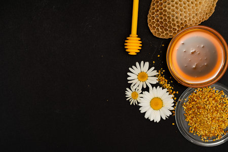 A yellow spoon of honey dipped in a jar of honey and bee bread scattered on a black background...