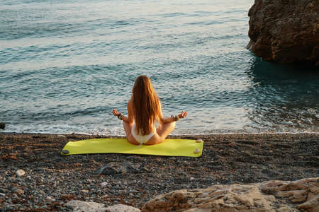 Beautiful woman in a bathing suit and long loose hair practicing yoga on the beach, Yoga Ardha Padmasana in the half-lotus position.の写真素材