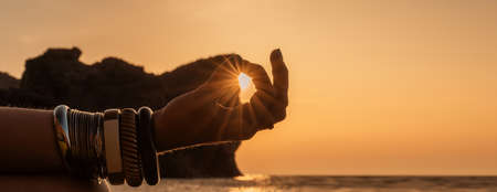 Banner. Young womans hand in bracelets. Practice yoga on the beach with sunset. Keeps fingers connected, the sun shines through them. The concept of a healthy lifestyle, harmony.の写真素材