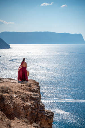 A girl with loose hair in a long red dress descends the stairs between the yellow rocks overlooking the sea. A rock can be seen in the sea. Sunny path on the sea from the rising sunの写真素材