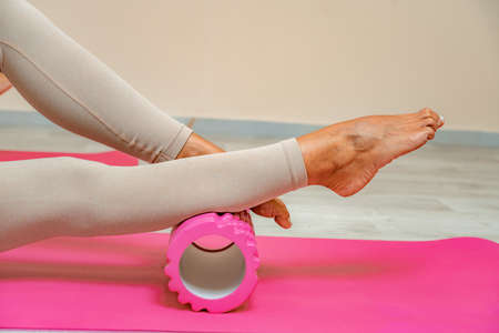 Fit athletic women in beige sportswear ride on a pink foam myofascial massage roller, massaging their muscles on the floor of a yoga mat in the gym. Active girls do a fitness workout in the gym. MFRの写真素材