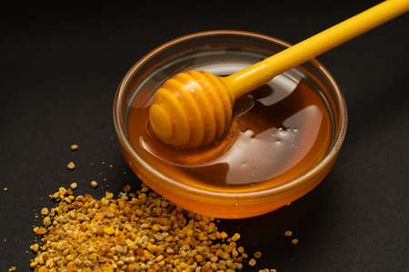 A yellow spoon of honey dipped in a jar of honey and bee bread scattered on a black background. Honey dripping around, nice and inviting photos. Healthy food concept.の写真素材