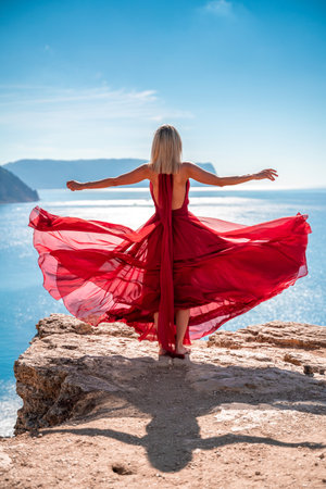 A girl with loose hair in a long red dress descends the stairs between the yellow rocks overlooking the sea. A rock can be seen in the sea. Sunny path on the sea from the rising sunの写真素材