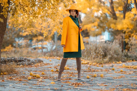 Beautiful woman walks outdoors in autumn. She is wearing a yellow coat, yellow hat and green dress. Young woman enjoying the autumn weather. Autumn contentの写真素材