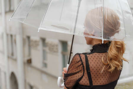The blonde stands under a transparent umbrella during the rain. The fall season. Rear view. The woman is dressed in a black lace dress, her hair pulled back in a ponytail.の写真素材