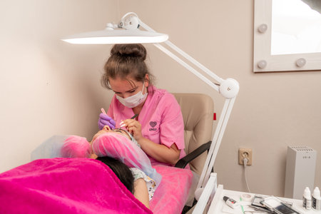Master lashmaker, diligently extends eyelashes to the client, on the couch, in the beauty salon. A...