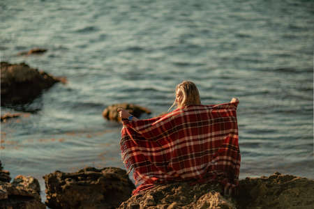 Attractive blonde Caucasian woman enjoying time on the beach at sunset, sitting in a blanket and looking to the side, with the sunset sky and sea in the background. Beach vacation.の写真素材