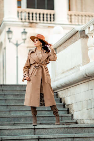 Outdoor fashion portrait of young elegant fashionable brunette woman, model in stylish hat, choker and light raincoat posing at sunset in European city.の写真素材