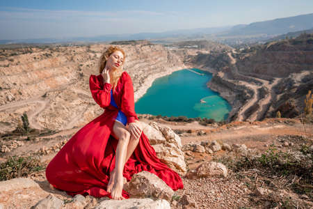 A beautiful girl in a red long dress, Sits on a rock high above the lake in the afternoon. Against the background of the blue sky and the lake in the shape of a heart.の写真素材