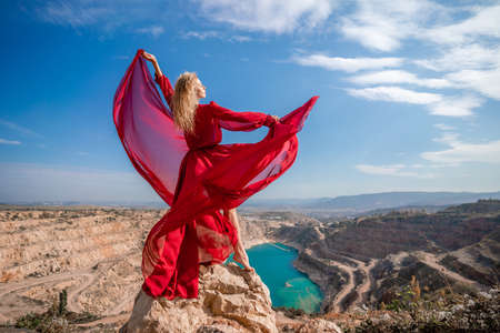 Side view of a beautiful sensual woman in a red long dress posing on a rock high above the lake in the afternoon. Against the background of the blue sky and the lake in the form of a heartの写真素材