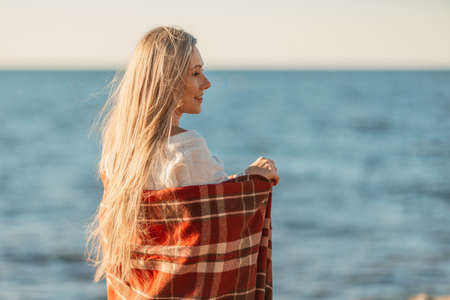 Attractive blonde Caucasian woman enjoying time on the beach at sunset, walking in a blanket and looking to the side, with the sunset sky and sea in the background. Beach vacationの写真素材