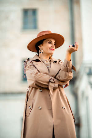 Outdoor fashion portrait of young elegant fashionable brunette woman, model in stylish hat, choker and light raincoat posing at sunset in European city.の写真素材