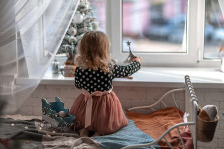 A little girl is sitting on the bed by the window and decorating a small tree with tiny Christmas toys. Happy healthy child celebrating a traditional family holiday. Adorable baby.の写真素材