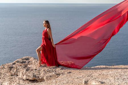 A girl with loose hair in a red dress stands on a rock rock above the sea. In the background, the sea and the rocks. The concept of travelの写真素材