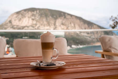 A glass of Latte Macchiato on the terrace of a summer cafe with views of the landscape, mountains and sea, a relaxed morning on vacationの写真素材