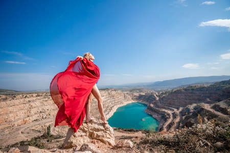Side view of a beautiful sensual woman in a red long dress posing on a rock high above the lake in the afternoon. Against the background of the blue sky and the lake in the form of a heartの写真素材