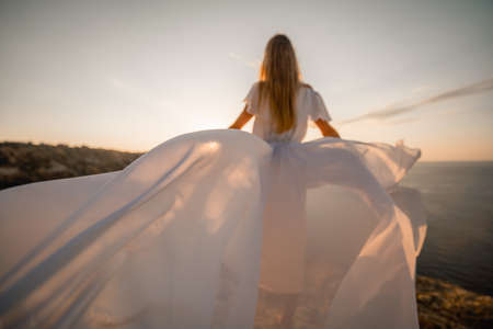 A beautiful young woman in a white light dress with long legs stands on the edge of a cliff above the sea waving a white long dress, against the background of the blue sky and the sea.の写真素材