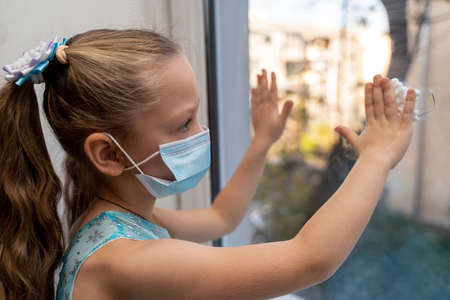 Girl child hygienic mask stay isolation at home for self quarantine for protect against virus  and air pollution. Little girls look out the window from the bedroom at homeの写真素材