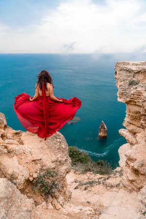 A girl with loose hair in a long red dress descends the stairs between the yellow rocks overlooking the sea. A rock can be seen in the sea. Sunny path on the sea from the rising sunの写真素材