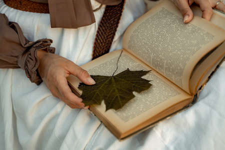 A woman in a white skirt and brown blouse sits and holds an open book in her hands. She is reading a book. There is a maple leaf in the knit as a bookmark.の写真素材