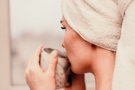 Young serene relaxed woman in spa bath towel drinking hot beverage tea coffee after taking shower...