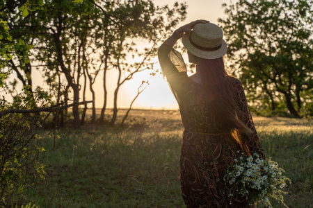 A happy woman travels alone in the forest at sunset, in a long dress, hat and a bouquet of flowers. Happy vacation. Rear view of a happy womanの写真素材