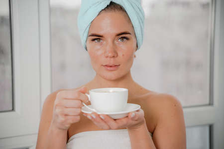 Young serene relaxed woman in spa bath towel drinking hot beverage tea coffee after taking shower bath at home. Beauty treatment, hydration concept.の写真素材
