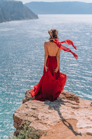 A girl with loose hair in a red dress waved her skirt on the yellow rocks overlooking the sea. Sunny path on the sea from the rising sun.の写真素材
