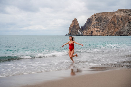 A beautiful and sexy brunette in a red swimsuit on a pebble beach, Running along the shore in the foam of the wavesの写真素材