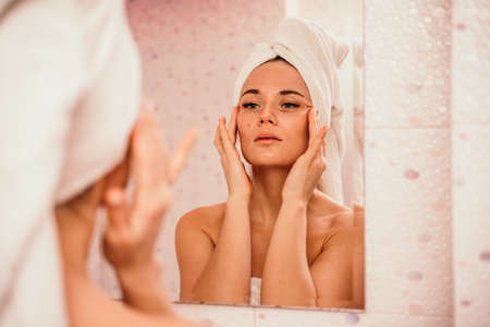 Young beautiful woman using face cream moisturizing lotion after bath. Pretty attractive girl in a towel on her head stands in front of a mirror in a home bathroom. Daily hygiene and skin careの写真素材