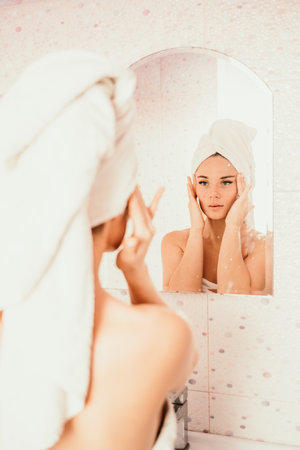 Young beautiful woman using face cream moisturizing lotion after bath. Pretty attractive girl in a towel on her head stands in front of a mirror in a home bathroom. Daily hygiene and skin careの写真素材