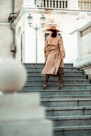 Outdoor fashion portrait of young elegant fashionable brunette woman, model in stylish hat, choker and light raincoat posing at sunset in European city.の写真素材