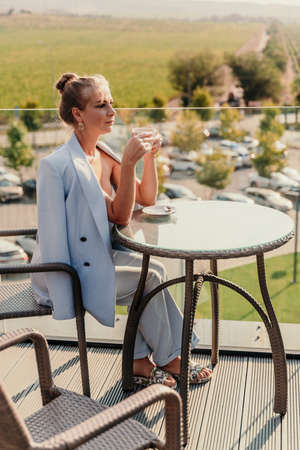 A middle-aged woman sits in a street cafe overlooking the mountains at sunset. She is dressed in a blue jacket and drinks coffee admiring the nature. Travel and vacation concept.の写真素材