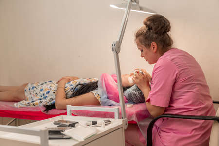 Master lashmaker, diligently extends eyelashes to the client, on the couch, in the beauty salon. A beauty master, a girl of 30 years old, European appearance, at work, in a bright roomの写真素材