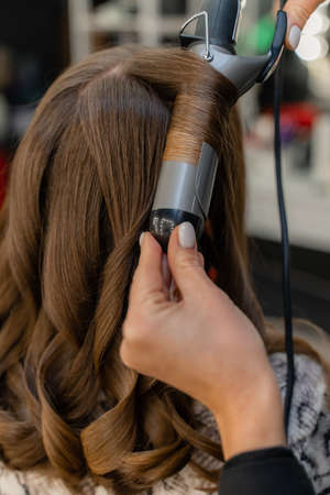 The hairdresser prepares a woman, makes a hairstyle with curls using a curling iron. Long light brown natural hair. Self care concept.の写真素材