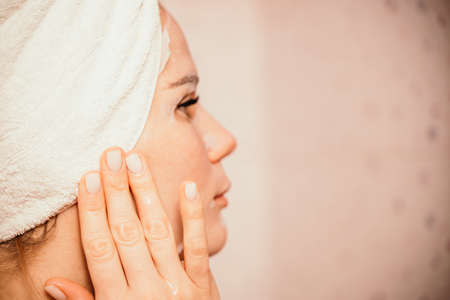 Young beautiful woman using a moisturizing facial mask after taking a bath. Pretty attractive girl in a towel on her head stands in front of a mirror in a home bathroom. Daily hygiene and skin careの写真素材