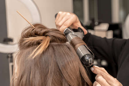 The hairdresser prepares a woman, makes a hairstyle with curls using a curling iron. Long light brown natural hair. Self care concept.の写真素材