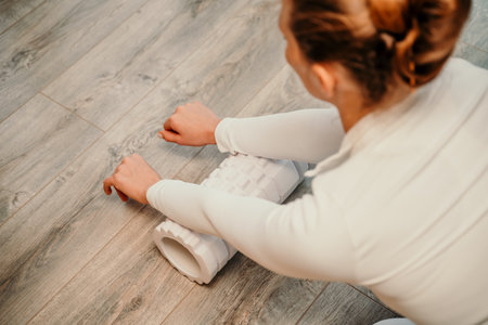 Adult athletic woman, in white bodysuit, performing fascia exercises on the floor - caucasian woman using a massage foam roller - a tool to relieve tension in the back and relieve muscle pain - the concept of physiotherapy and stretching training.の写真素材