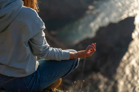 Woman tourist enjoying the sunset over the sea mountain landscape. Sits outdoors on a rock above the sea. She is wearing jeans and a blue hoodie. Healthy lifestyle, harmony and meditationの写真素材