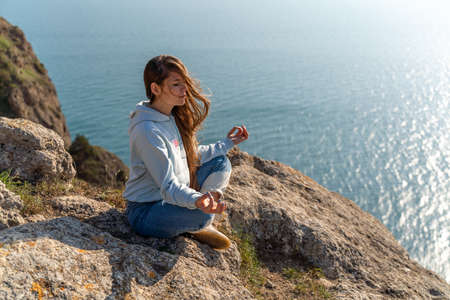 Woman tourist enjoying the sunset over the sea mountain landscape. Sits outdoors on a rock above the sea. She is wearing jeans and a blue hoodie. Healthy lifestyle, harmony and meditationの写真素材
