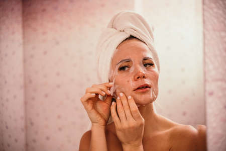 Young beautiful woman using a moisturizing facial mask after taking a bath. Pretty attractive girl in a towel on her head stands in front of a mirror in a home bathroom. Daily hygiene and skin careの写真素材