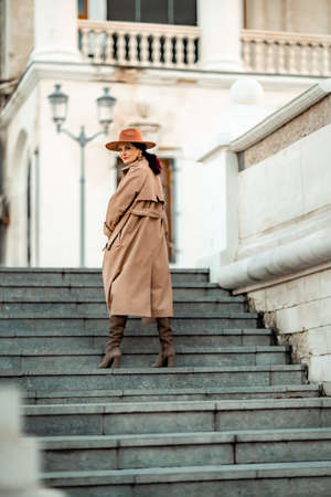 Outdoor fashion portrait of young elegant fashionable brunette woman, model in stylish hat, choker and light raincoat posing at sunset in European city.の写真素材