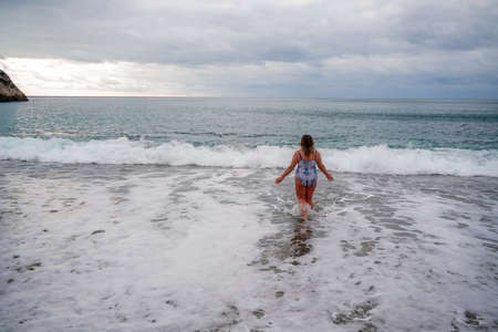 A plump woman in a bathing suit enters the water during the surf. Alone on the beach, Gray sky in the clouds, swimming in winter.の写真素材