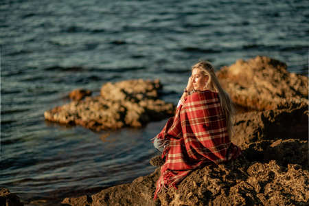 Attractive blonde Caucasian woman enjoying time on the beach at sunset, sitting in a blanket and looking to the side, with the sunset sky and sea in the background. Beach vacation.の写真素材